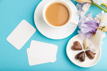 White business card with cup of cioffee, chocolate candies and iris flowers on blue pastel background. top view, copy space.