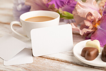Obraz na płótnie Canvas White business card with cup of cioffee, chocolate candies and iris flowers on white wooden background. side view, copy space.