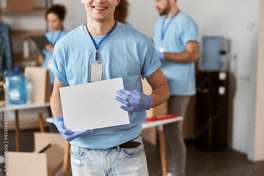 Wall mural Cropped shot of caucasian young male volunteer in blue uniform and protective gloves smiling at camera, holding blank white paper banner for your ad - Wall murals
