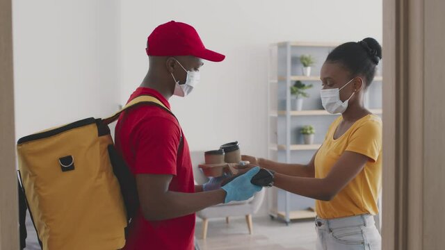 African american delivery man wearing protective mask and gloves giving coffee to lady, black woman using smartwatch