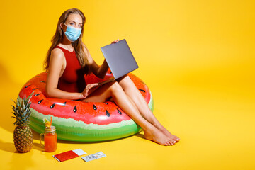  young woman dressed in red swimsuit sitting on inflatable circle on the yellow background. wearing a mask with a laptop near the cocktail pineapple tickets looking at the camera 