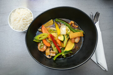 Vegetables and shrimps in teriyaki sauce in a black bowl served with rice on a gray table, high angel view from above, selected focus