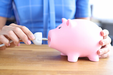 Woman is holding electrical plug and and pink piggy bank closeup