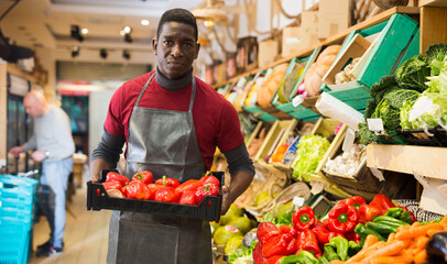 Obraz premium Focused African man working at farmer market, holding box with fresh organic red sweet peppers ..