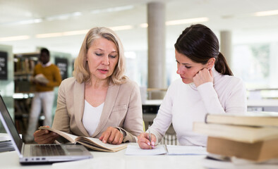 Friendly female tutor helping to diligent positive girl preparing for exam in library. High quality photo