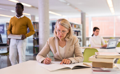 Elderly woman makes notes in notebook and read book in public library. High quality photo