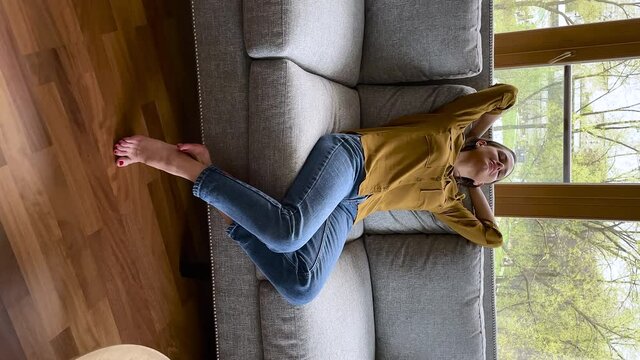 Vertical view woman relaxing on sofa at modern home. Carefree female in casual clothes stretching leaned on couch cushion enjoy day off alone breath fresh conditioned air. Stress free weekend concept