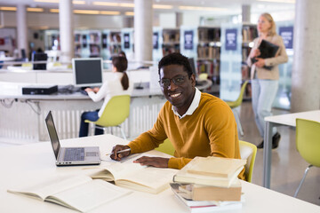 Portrait of smiling african-american male student with laptop and book in public library. High...