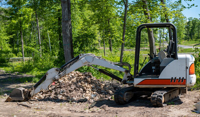 A mini excavator with hydraulic scoop sits on the dirt next to a pile of rocks at a new home construction lot, with trees in back of site.