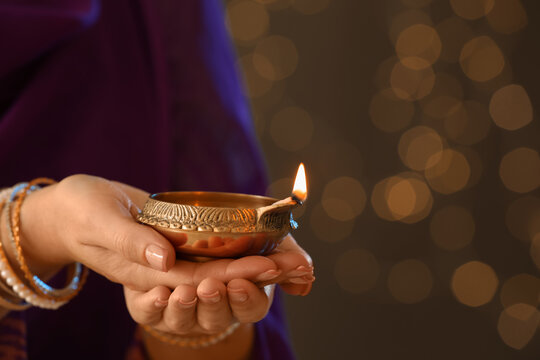 Woman holding lit diya lamp in hands against blurred lights, closeup. Diwali celebration