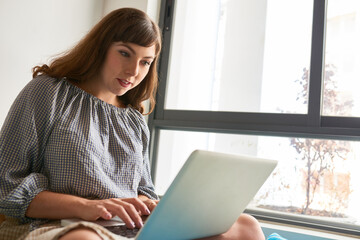 Positive woman sitting on window sill, coding or working with online documents