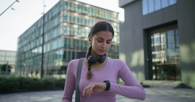 Time to start training. Calm pensive young woman in sportswear checks time on smartwatch has scheduled workout with coach carries necessary things goes away to sports gym maintains vitality.