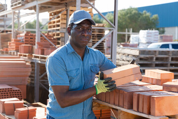 African-american male worker carrying clay bricks in outdoor construction material storage.