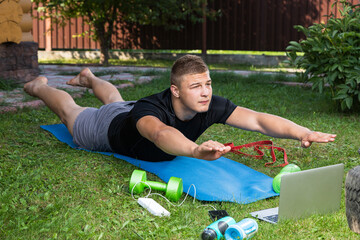 The young man goes in for sports in park. Sportsman with blond hair makes a plank, watches a movie and studies from a laptop  on carpet in  backyard  in summer day