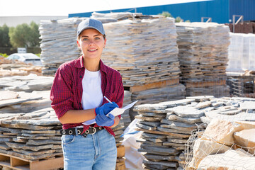 Young attractive female worker with papers checking quantity of natural stone tiles in warehouse of...