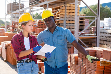 African-american man and European woman standing beside clay brick stacks in outdoor construction material storage. Man carrying flat clay brick in hands.