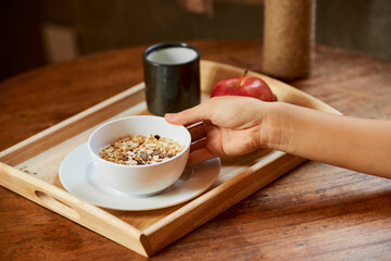 Woman putting cup of muesli with nuts and raisins on wooden tray when preparing healthy breakfast