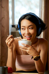 Portrait of joyful young woman listening to music in headphone and eating delicious granola with yogurt