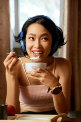 Pretty attractive young smiling woman listening to good music in headphone and eating muesli with...