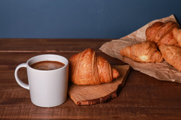 Fresh baked croissants with craft paper, chopping board, mug of black coffee on wooden table. Cup of hot drink, buns in wrap. Food, breakfast, bakery shop, morning menu, cafe concept. Side view