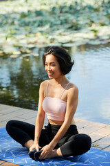 Fototapeta na wymiar Happy slim fit young woman sitting on yoga mat in lotus position on wooden pier