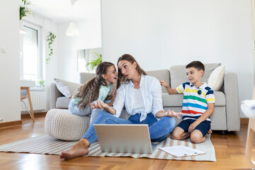 Small mischievous kids noising and distracting mother freelancer trying to concentrate on laptop and squeezing head working at table in light living room