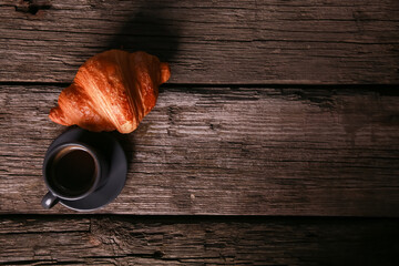Expresso cup and croissant on shabby rustic wooden table background. Fresh baked bun and hot black...
