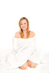 Fototapeta na wymiar Smiling young woman sitting on her bed covering herself with bedclothes, isolated on white background