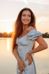 Fashion portrait of charming young woman in blue dress posing by sunset landscape and looking away  - 455220739
