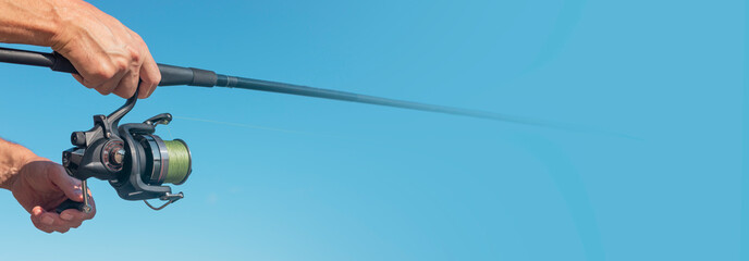 Fishing rod reel in fisherman hands over clear blue sky with copy space for text. Horizontal banner.
