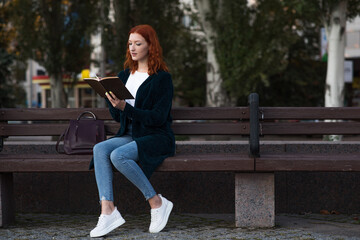 A young and attractive redhead Caucasian girl is reading a book while sitting on a bench. Education concept.