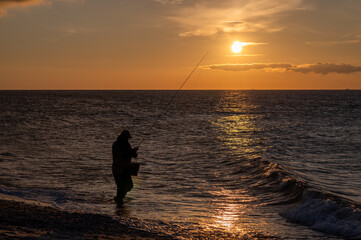 Angler steht mit seiner Angel am Meer im Abendrot bei Sonnenuntergang und angelt.