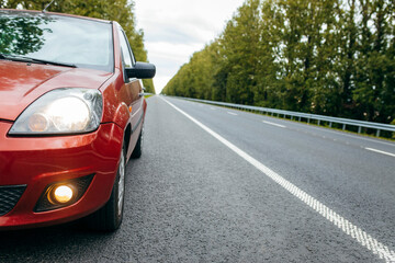 Fototapeta premium Red car with low beam on, parked on road background nature green trees