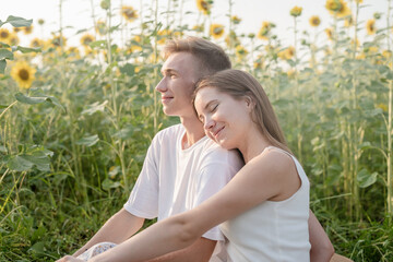 Fototapeta premium Young couple having picnic on sunflower field at sunset, woman lying on boyfriend shoulder
