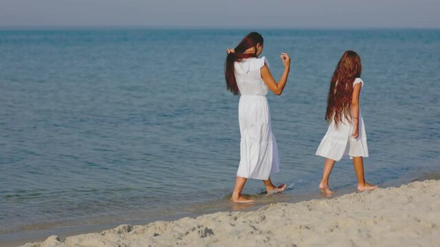 mom and daughter in white dresses are walking near the sea