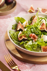 A beige colored plate with arugula salad with mozzarella, figs and pork meat jamon and balsamico dressing on a pink marble surface