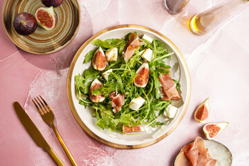 A beige colored plate with arugula salad with mozzarella, figs and pork meat jamon and balsamico dressing on a pink marble surface, top view