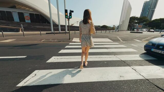 Back of stylish girl walking across pedestrian crossing in sunny day. Beautiful tourist young woman in short tender dress walks in city center.