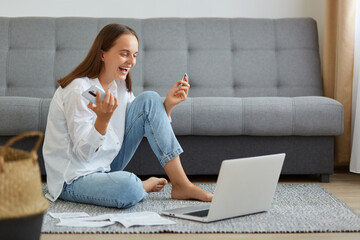 Portrait of happy woman wearing white shirt and jeans sitting on floor and looking at laptop...