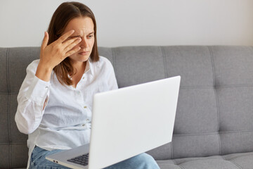 Portrait of unhappy tired female wearing white shirt and jeans sitting on sofa at home, holding notebook and looking at computer screen, working long hours, feels hurt in eyes, rubbers eye.