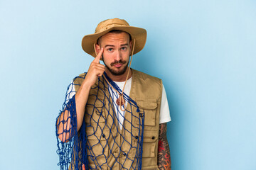 Young caucasian fisherman with tattoos holding net isolated on blue background  pointing temple with finger, thinking, focused on a task.