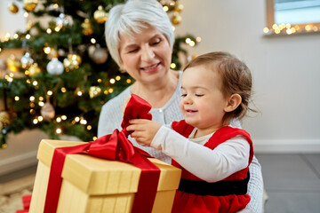 Fototapeta na wymiar christmas, holidays and family concept - happy grandmother and baby granddaughter opening gift box at home