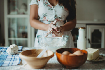 Lady wearing white apron molding dough.Baker at home.Messy table with baking ingredients.Milk bottle and eggs in bowl.Round doughs on blue and white checkered table cloth.