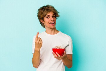 Young caucasian man with makeup a cereal bowl isolated on blue background  pointing with finger at you as if inviting come closer.