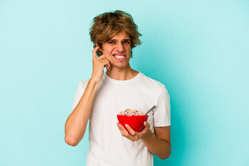 Young caucasian man with makeup a cereal bowl isolated on blue background  covering ears with hands.