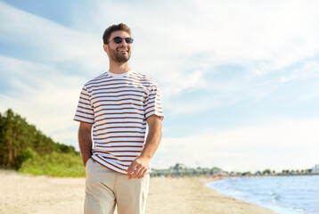 summer holidays and people concept - portrait of young man in sunglasses walking along beach in...