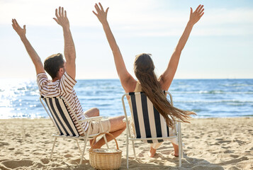 leisure, relationships and people concept - happy couple with picnic basket sitting in folding chairs with raised hands on summer beach