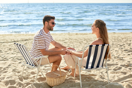 leisure, relationships and people concept - happy couple with picnic basket sitting in folding chairs and holding hands on summer beach