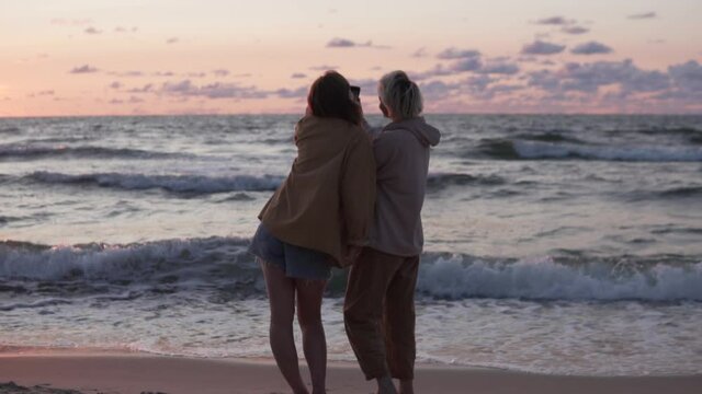 Two women friends take a photo on the beach at sunset, a view from the back. Romantic walk of two Lisbians.