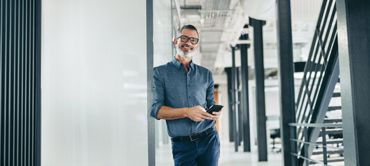 Smiling mature businessman holding a smartphone in an office - Powered by Adobe